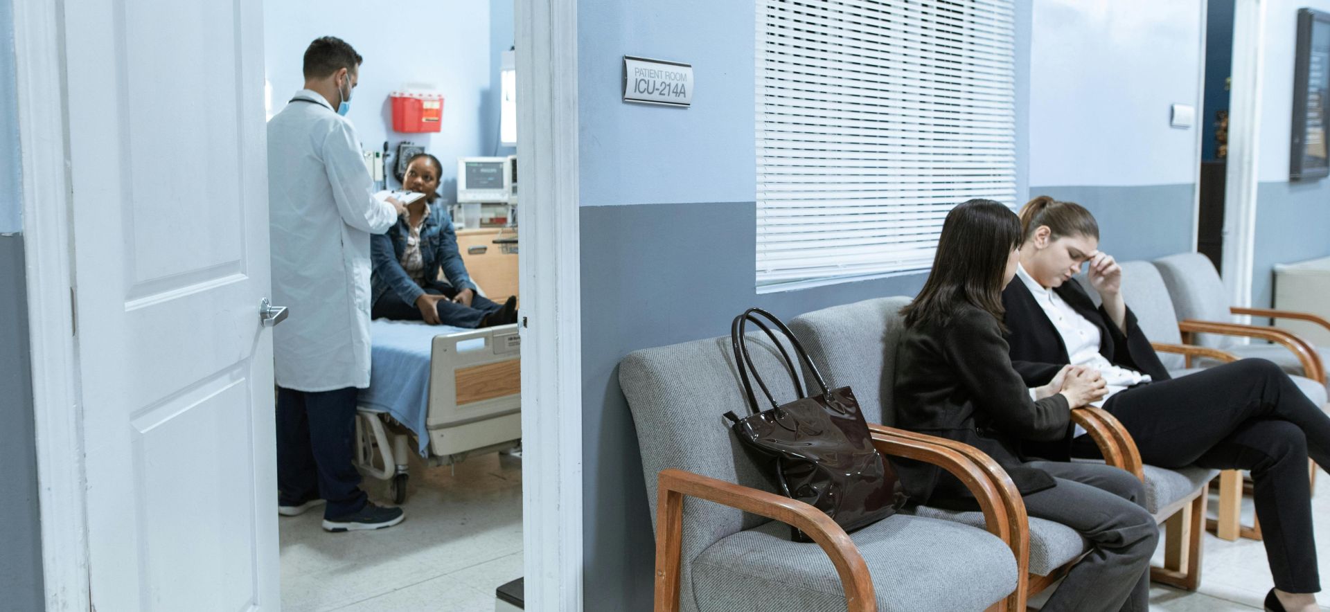 Doctor with patient in ICU, women in waiting area showing concern.